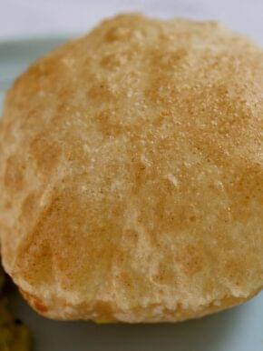 Golden brown, puffy poori bread freshly fried to perfection. One large, round puri is the focus, showing its layered, flaky texture and subtle speckles from frying. Other small pooris are stacked in the background. This popular Indian bread is fried to form air pockets, creating a crisp exterior and soft, pillowy interior.